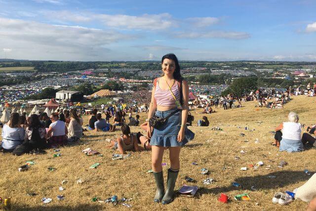 A woman posing for a picture on a hill with views of Glastonbury Festival in the background.