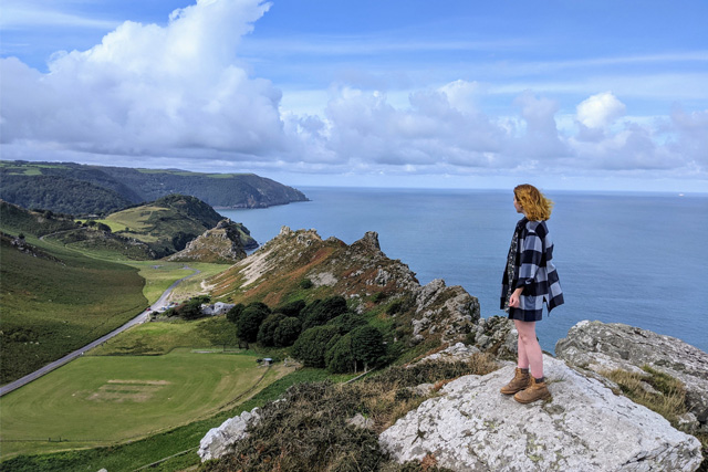 Open view of rock formations at the Valley of Rocks and the sea behind, with a lady stood at a viewpoint in the foreground.